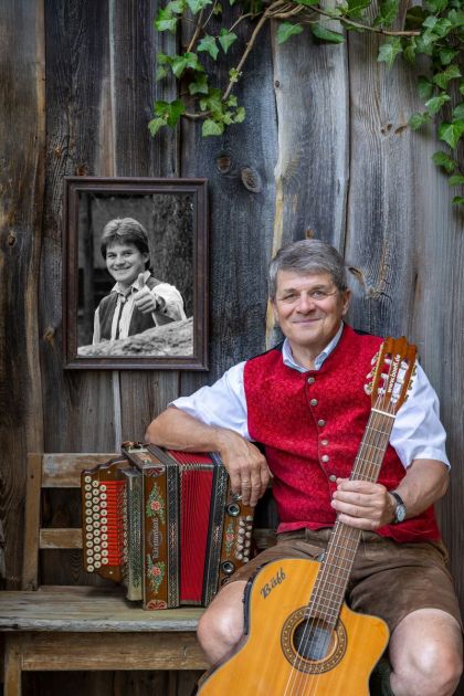 Josef Bäff Piendl sitzt mit Gitarre und Harmonika auf einer Holzbank