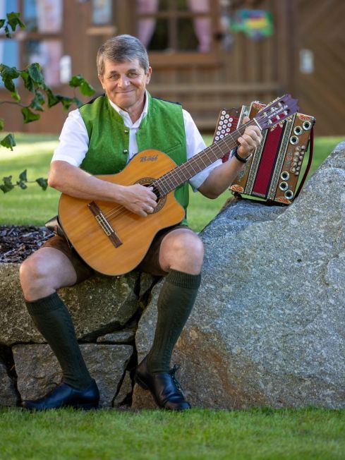 Bäff spielt sitzend auf einem Stein mit der Gitarre, dahinter steht seine Harmonika